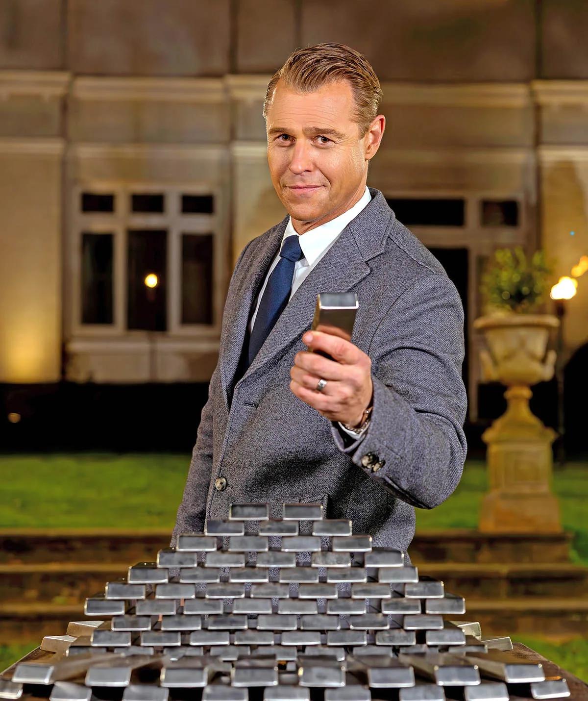 Rodger Corser in a classy grew suit in front of a mansion holding a silver bar he has just picked up from a pyramid of bars in a promotional shoot for The Traitors