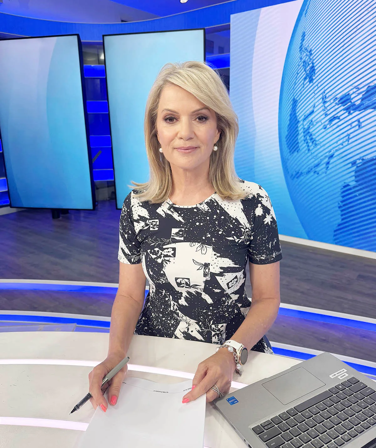 Sandra, wearing black and white, sits behind the newsdesk. 