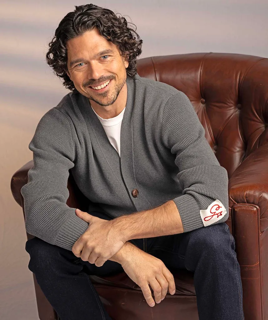 Luke sits on a brown leather chair and is all smiles with his brown curls and grey cardigan