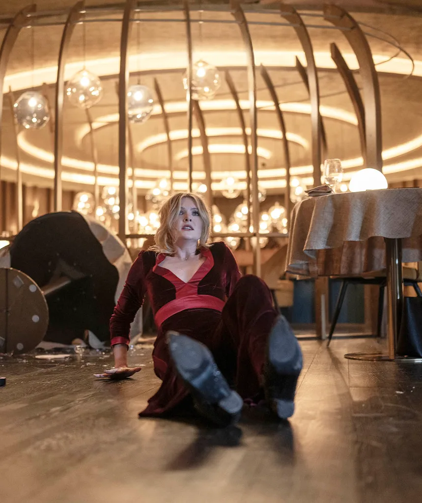 Meghann on the floor with tables around her tipped over and smashed wine glasses in a fancy restaurant with a look of terror on her face