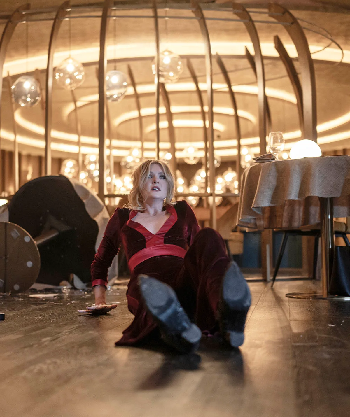 Meghann on the floor with tables around her tipped over and smashed wine glasses in a fancy restaurant with a look of terror on her face
