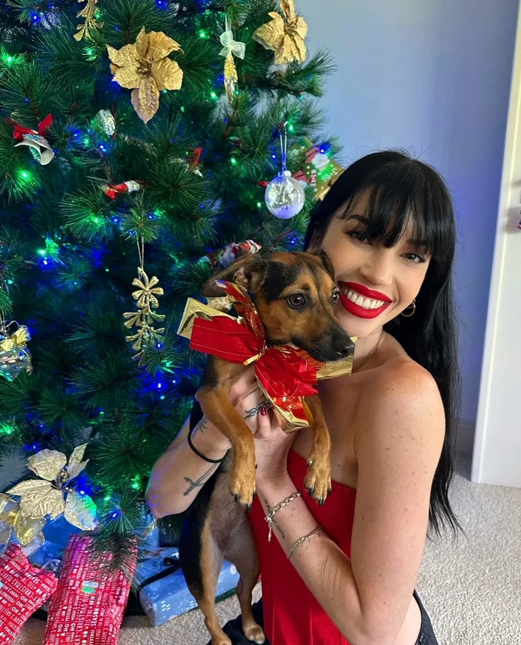 Shannon Clarke with her dachshund, Chilli,  wearing big red bow in front of a Christmas tree 