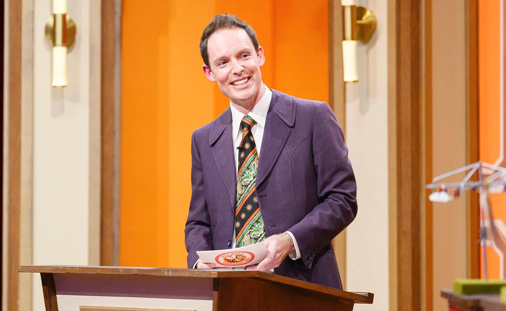 Guy Montgomery, in a purple jacket, stands at the podium on the set of his spelling bee show.