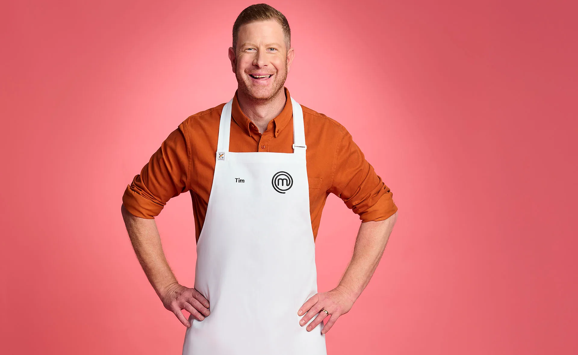 Tim stands in an orange tee with the white MasterChef apron on, hands on his hips, smiling against a pink backdrop