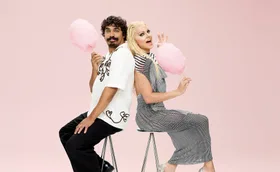 Tony in a white button up shirt and black pants sits back to back with Courtney in with white blonde hair and a silver dress as they both smile and hold sticks of fairy floss against a pink background