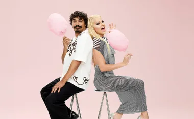 Tony in a white button up shirt and black pants sits back to back with Courtney in with white blonde hair and a silver dress as they both smile and hold sticks of fairy floss against a pink background