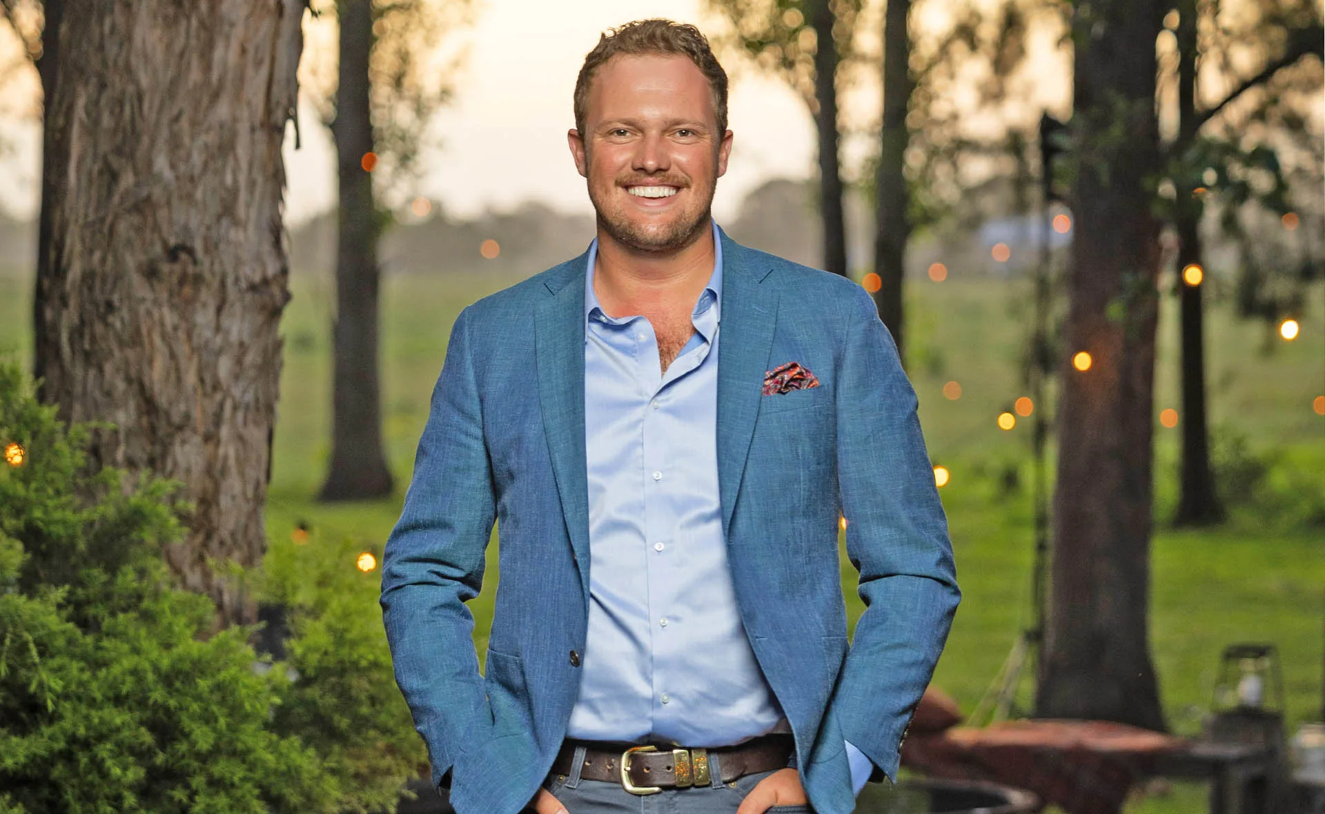 Corey, wearing a blue jacket, stands with his hands in his pockets, in front of some trees.