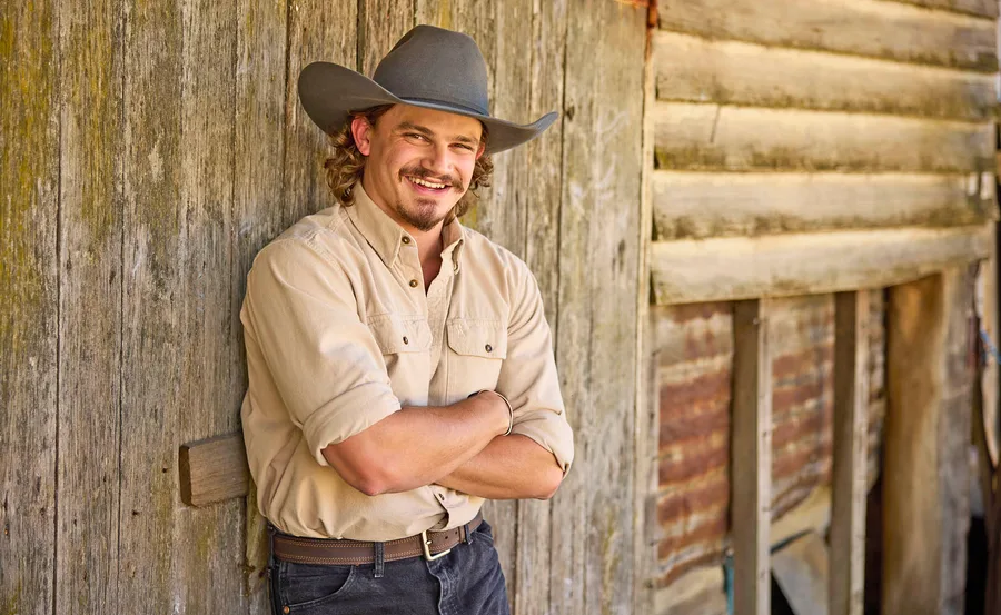 Jarrad, wearing a cowboy hat, leans against a wooden wall with his arms folded.