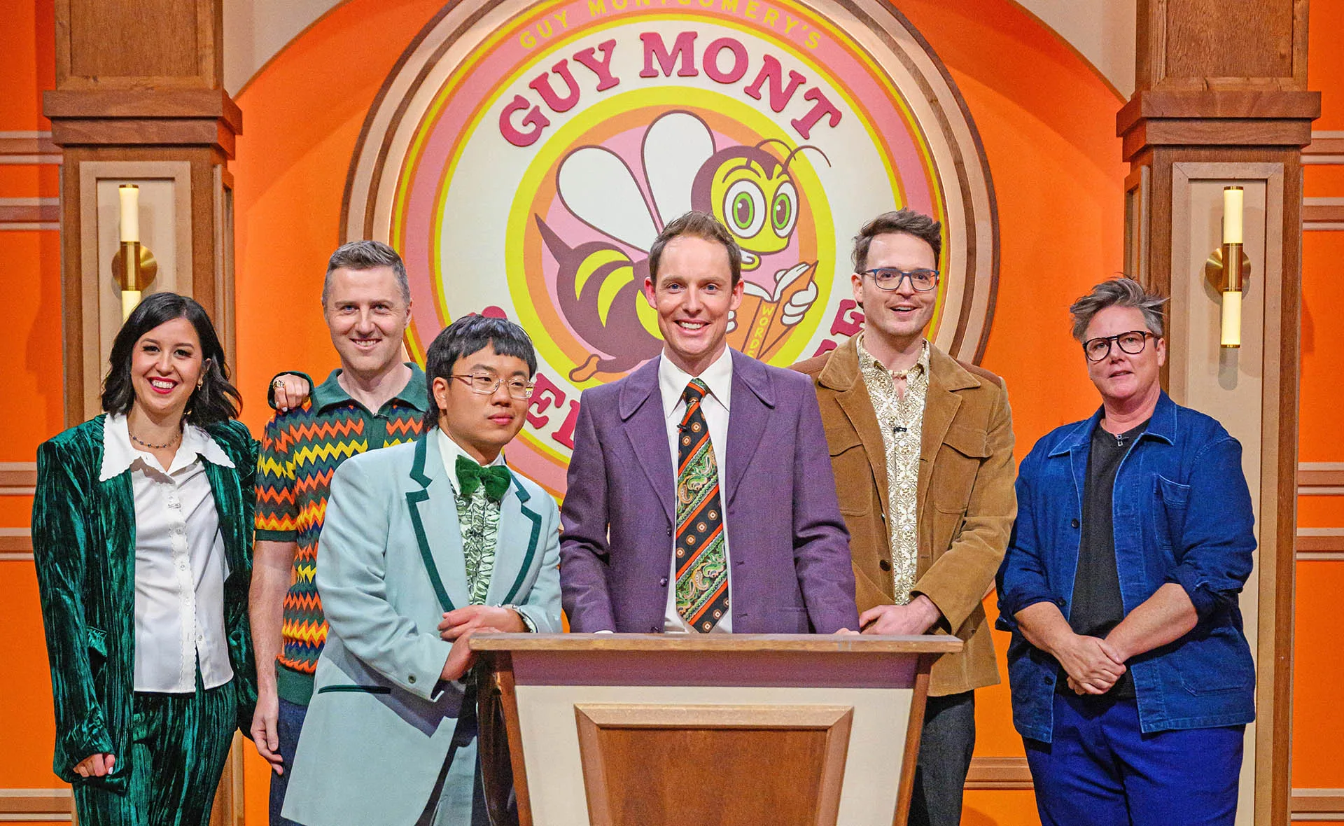 Guy Montgomery, in a purple jacket and a tie, stands behind a podium, with his sidekick Aaron Chen and competitors behind him.