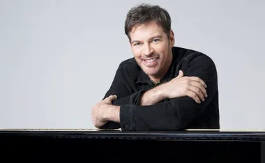 Harry Connick Jr, in black, leans forward with his elbows on a piano