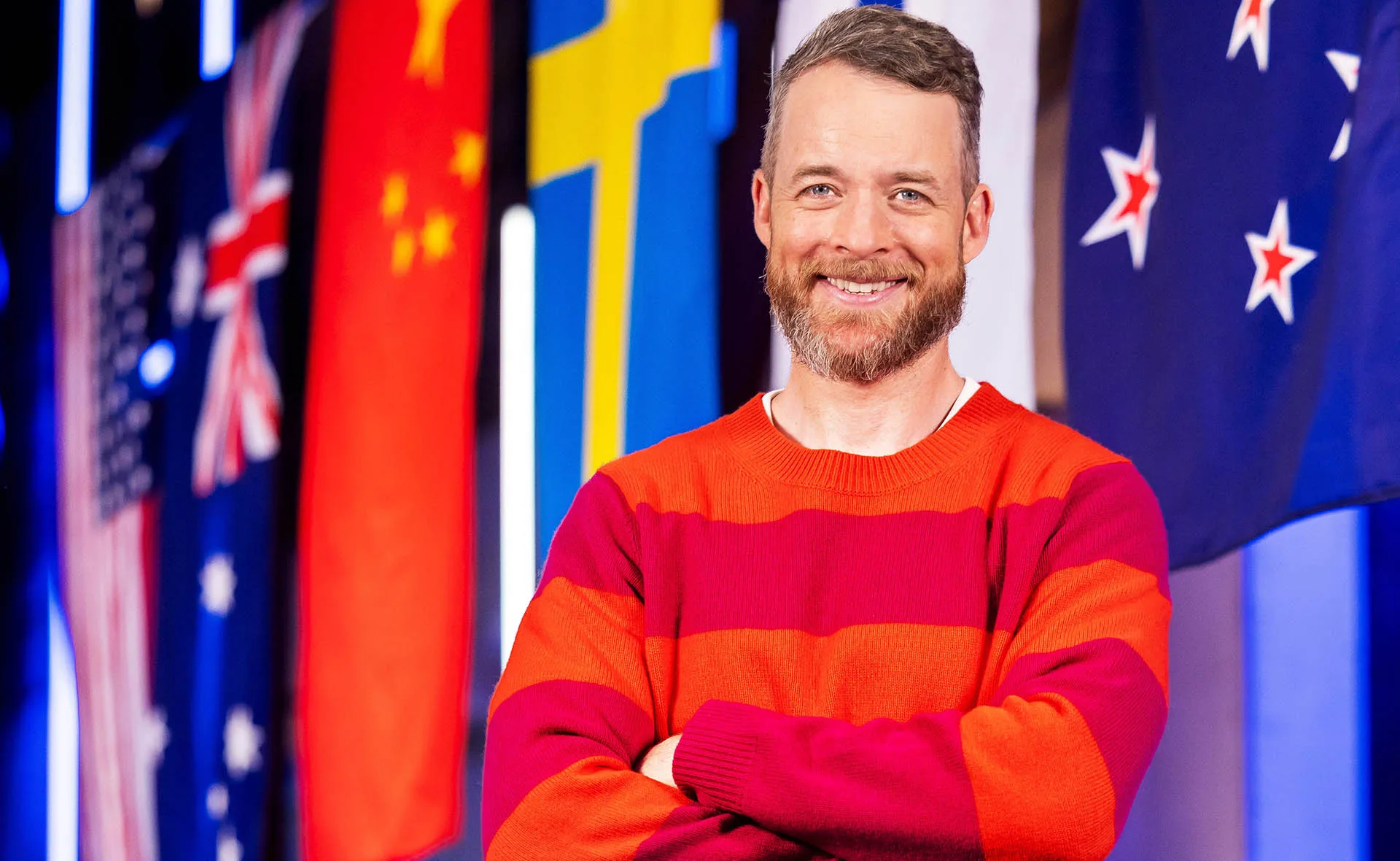 Hamish, in a striped red top, stands with his arms folded on the set of LEGO Masters, with flags behind him.