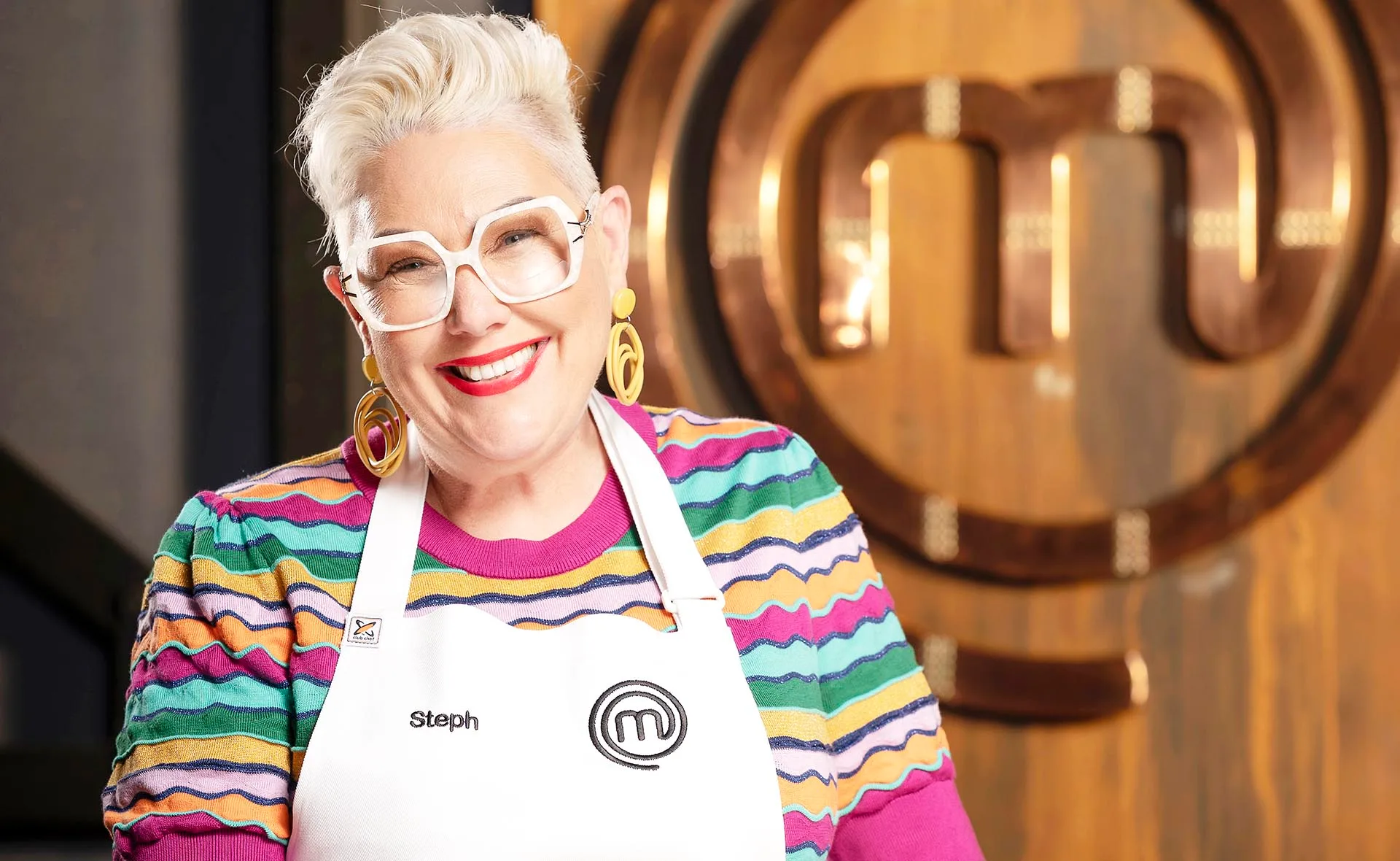 Steph pictured with short blonde hair, white glasses, yellow earrings, a striped shirt, MasterChef apron smiling