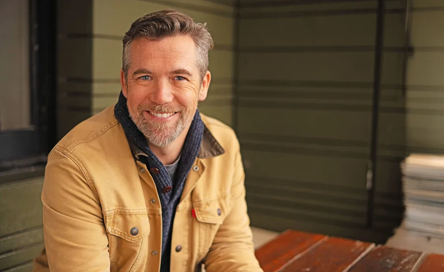 Patrick Brammall, wearing a brown jacket, smiles at the camera.