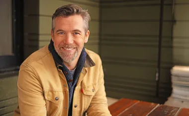 Patrick Brammall, wearing a brown jacket, smiles at the camera.