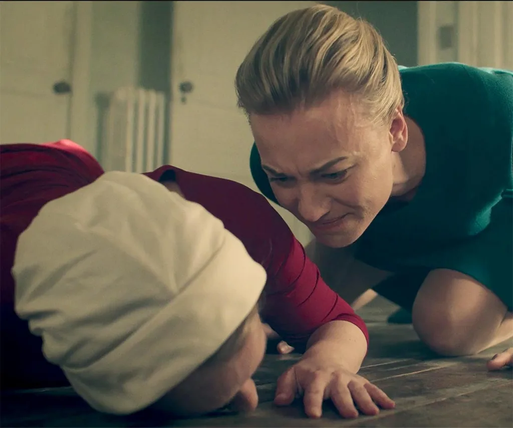 Serena cries and leans over June with angry tears in her face as June is collapsed on the floor in pain in her Handmaid's Tale outfit