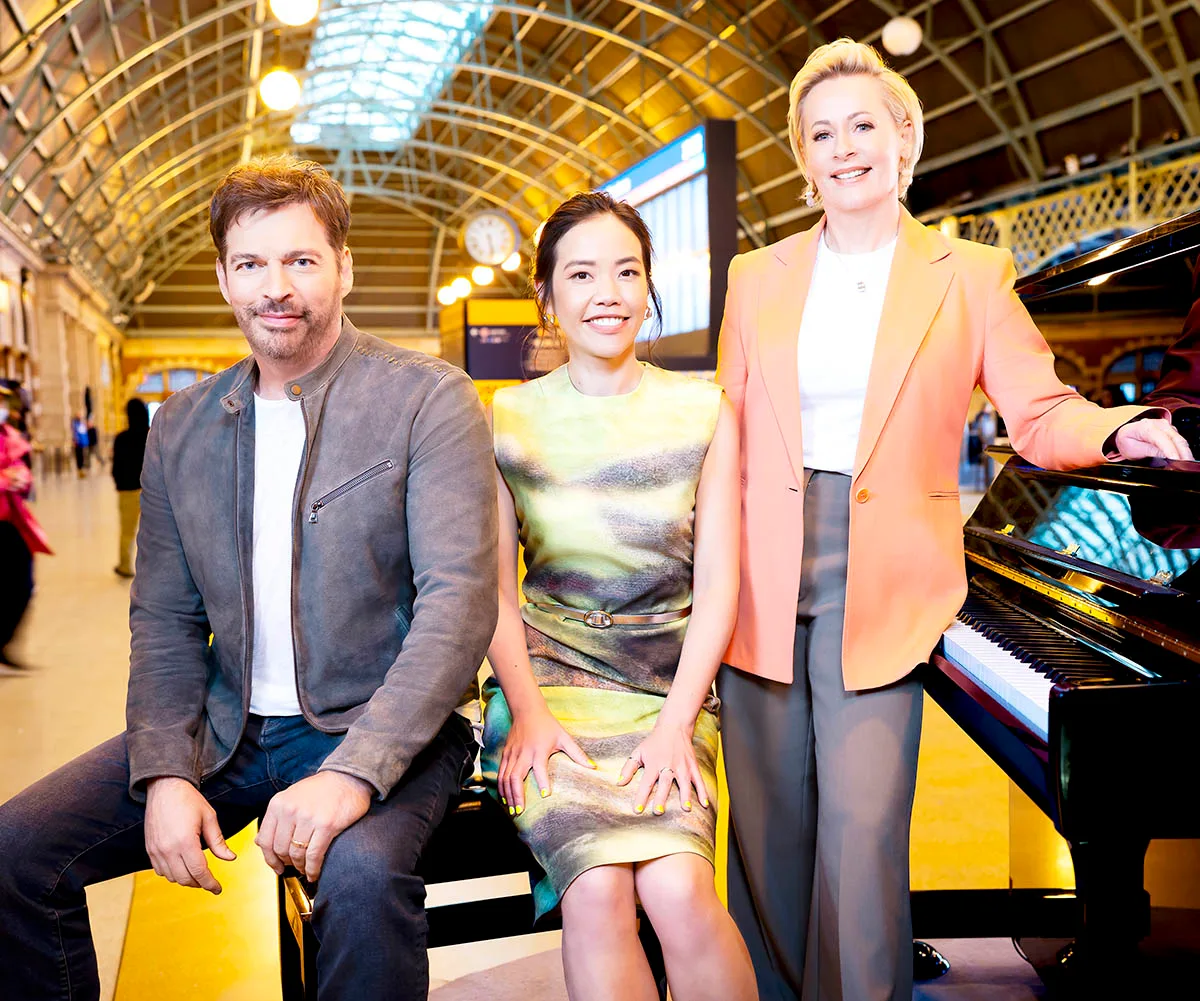Harry sits next to Andrea Lam while Amanda Keller stands next to them, her hand on a piano.