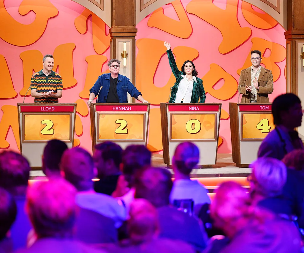 Four spelling bee contestants stand on stage. One has her hand in the air.