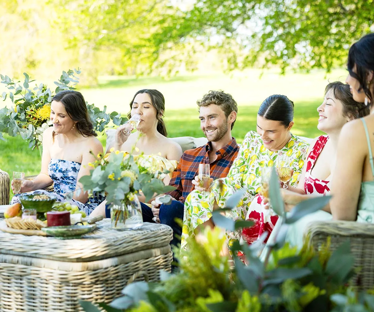 Farmer Jack sits on a seat in a garden on a sunny day, smiling, in the middle of a group of girls in dresses. 