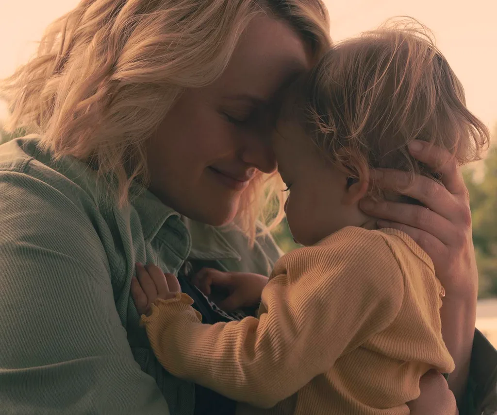 June in a denim jacket smiling as she holds her baby in a loving embrace on the train in new season of The Handmaid's Tale