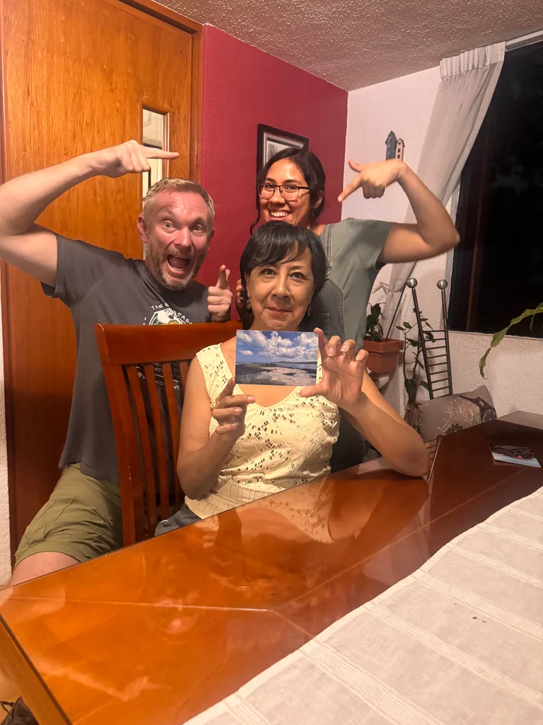 Jon Beardmore sitting in a dining room with mother and daughter pointing at the postcard Jon delivered