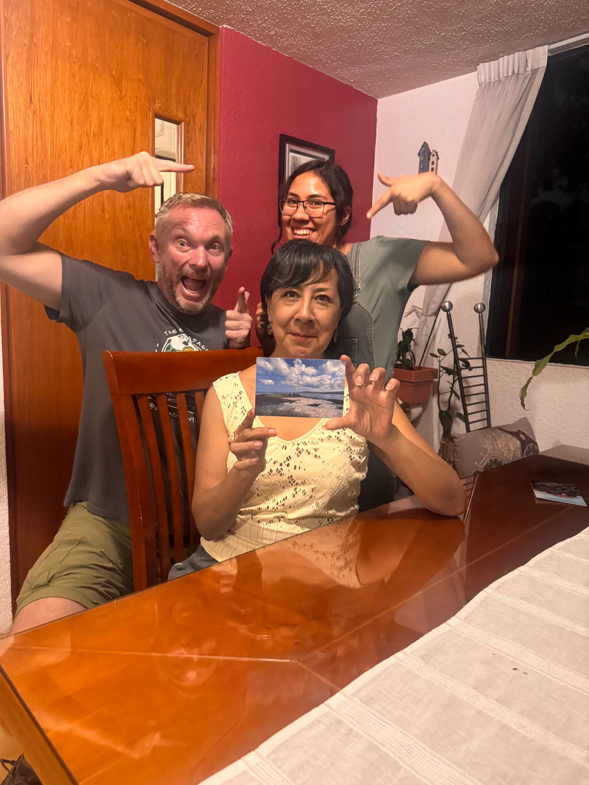Jon Beardmore sitting in a dining room with mother and daughter pointing at the postcard Jon delivered