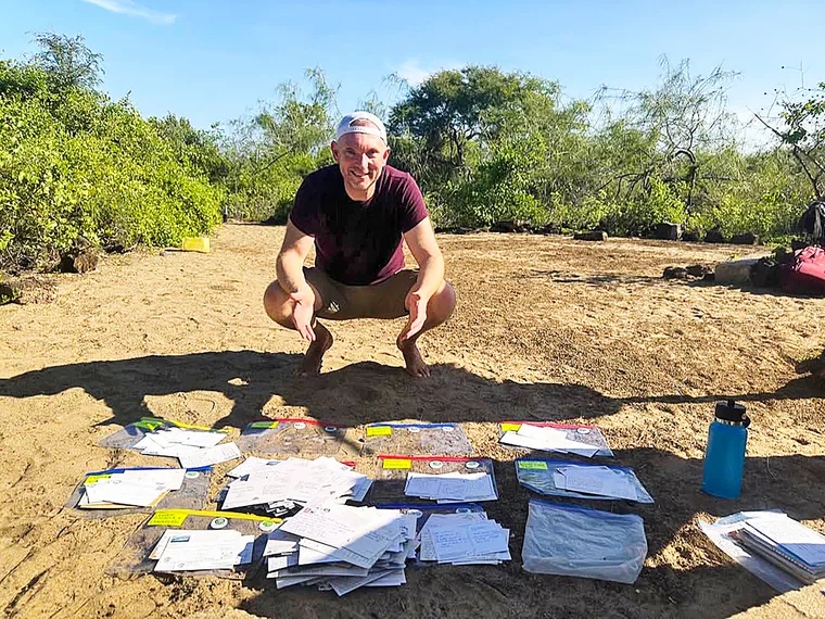 Jon Beardmore barefoot, squatting on Galapagos sand with parcels of mail on the ground in front of him
