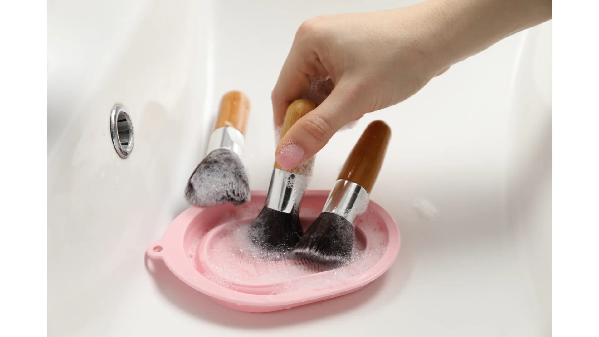 A woman's hand washes makeup brushes in a pink tray.