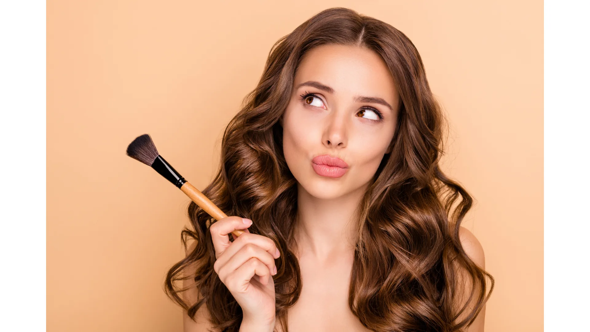 A brunette woman wearing makeup pouts and looks off to the side while holding a makeup brush.