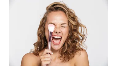 A woman laughs cheekily with her eyes closed while holding a makeup brush up to her face.