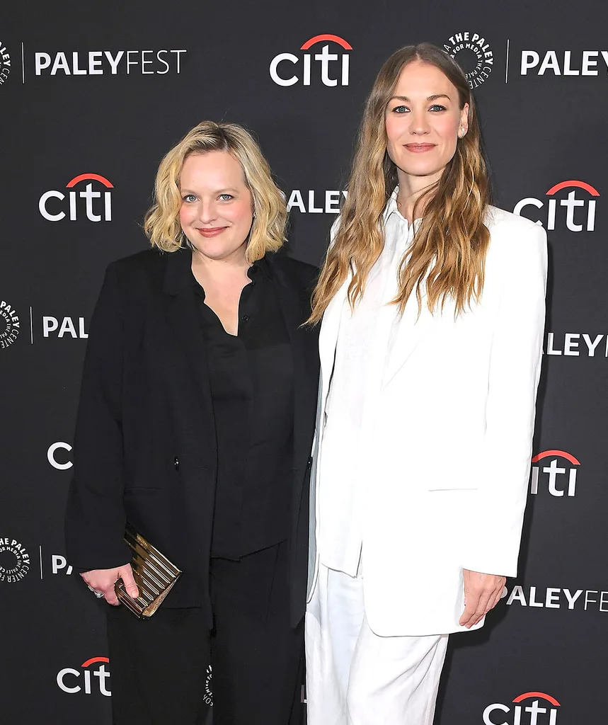 Elizabeth Moss in a black suit and Yvonne Strahovski in a white suit pictured against a media wall smiling at The Handmaid's Tale premiere
