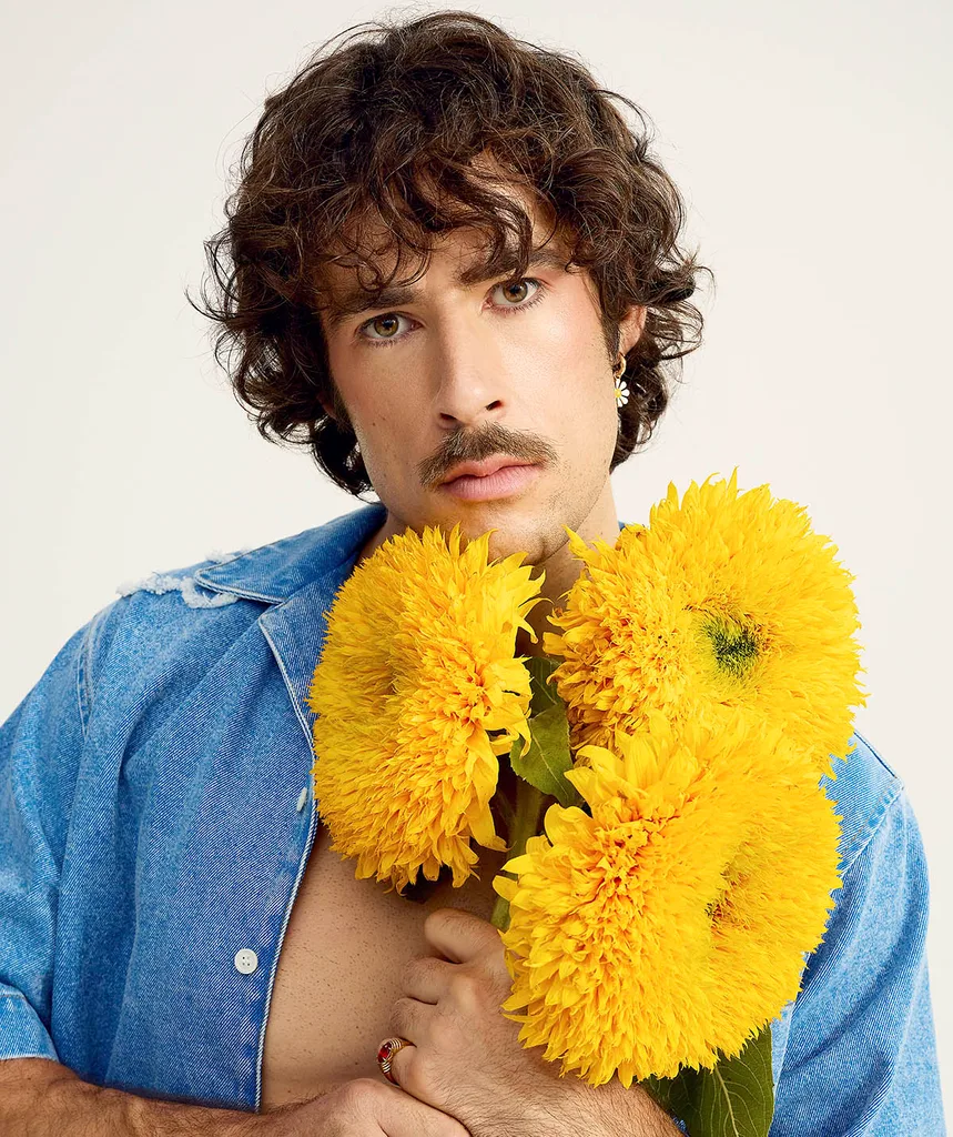 Performer Go-Jo pictured holding bright yellow flowers while pouting, wearing an open denim shirt with his signature curly brown locks.