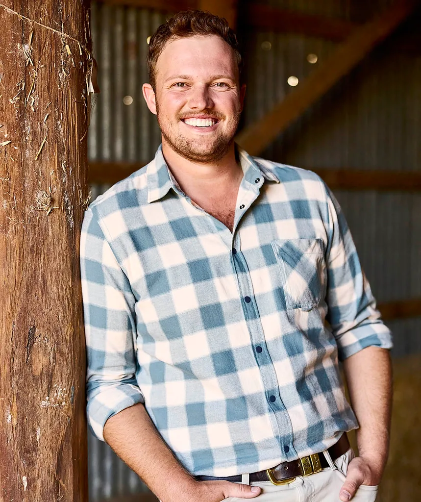 Farmer Corey, in a checked shirt, leans on a post inside a shed, smiling.