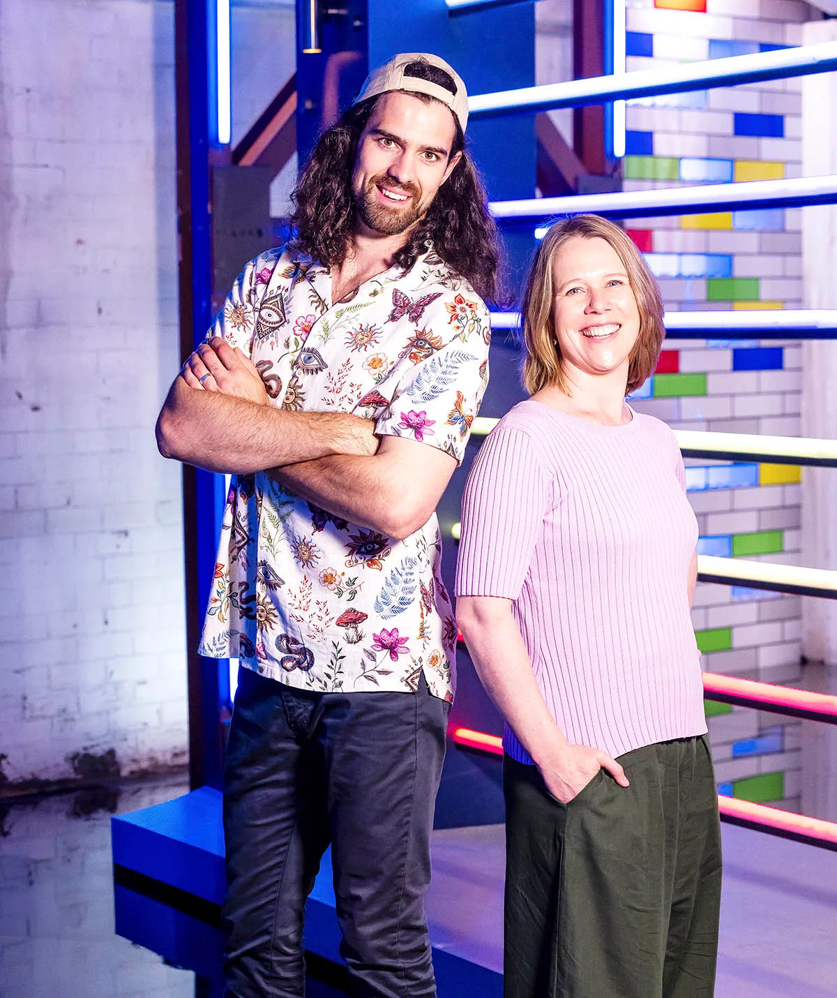 Left: Own with arms crossed long curly brown hair, party shirt and a backwards hat.
Right; Gabby in khaki pants, pink sweater shirt and short strawberry blonde hair. 
Both are smiling and on the LM set.