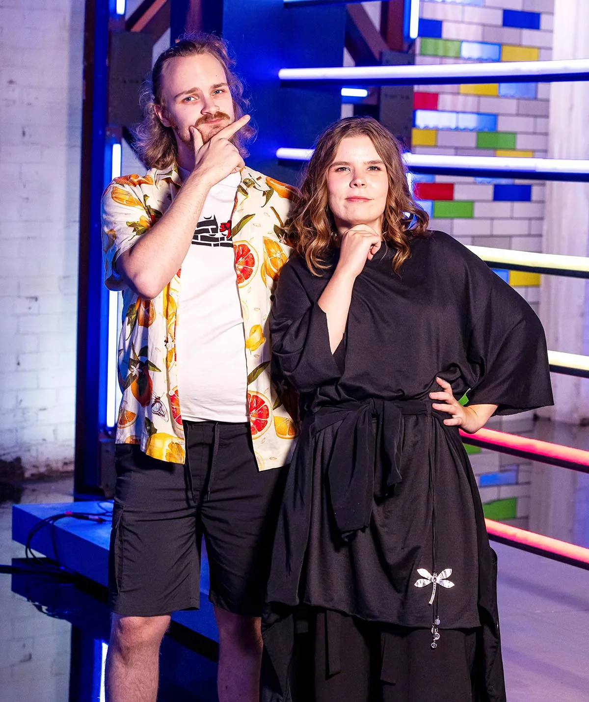 On left: Oskari in black shorts, white t-shirt, open colourful button up over the top as he pulls a silly face with half his blonde hair tied up in a bun. 
On right: Aura in a long black dress with medium length brown hair and her first placed under her chin on LM set.