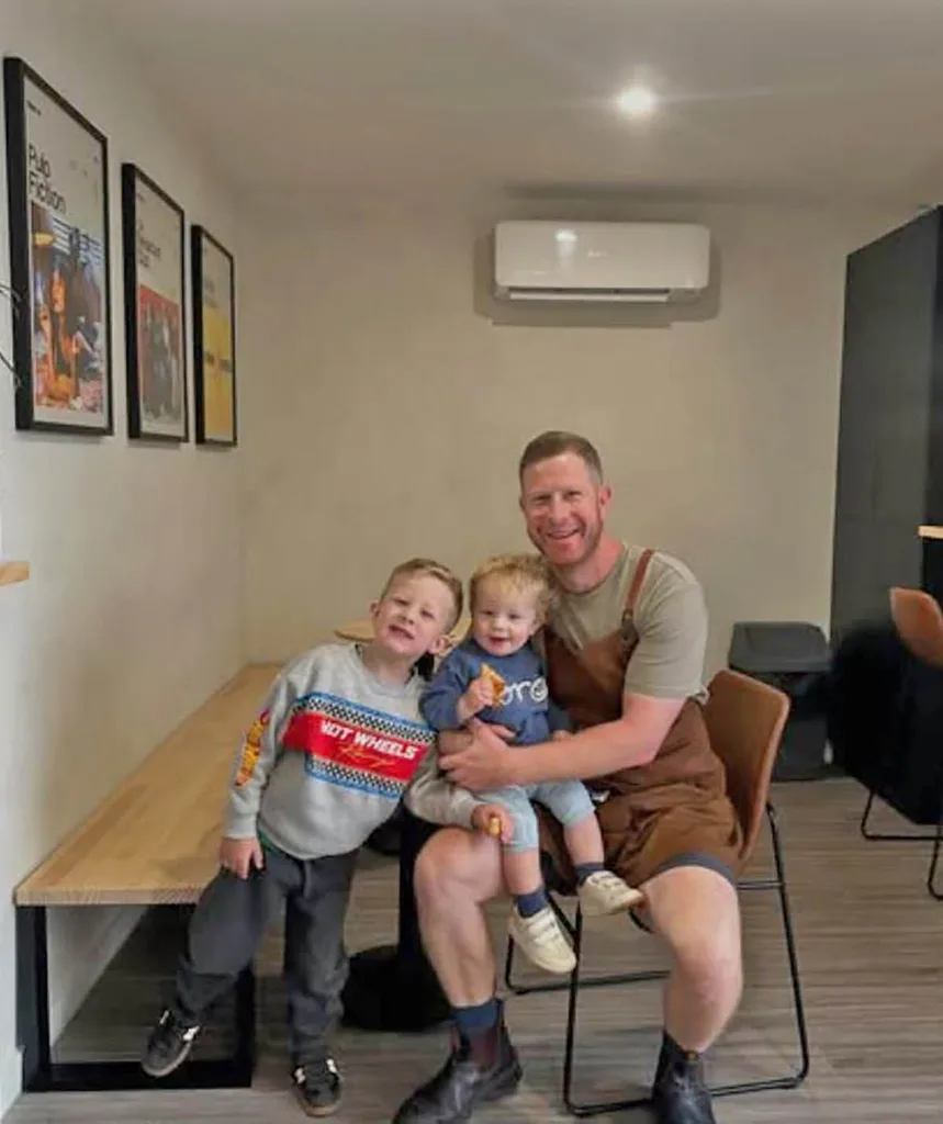 Tim sits smiling with his two sons, Toby and Jude as he wears a grey t-shirt and brown apron