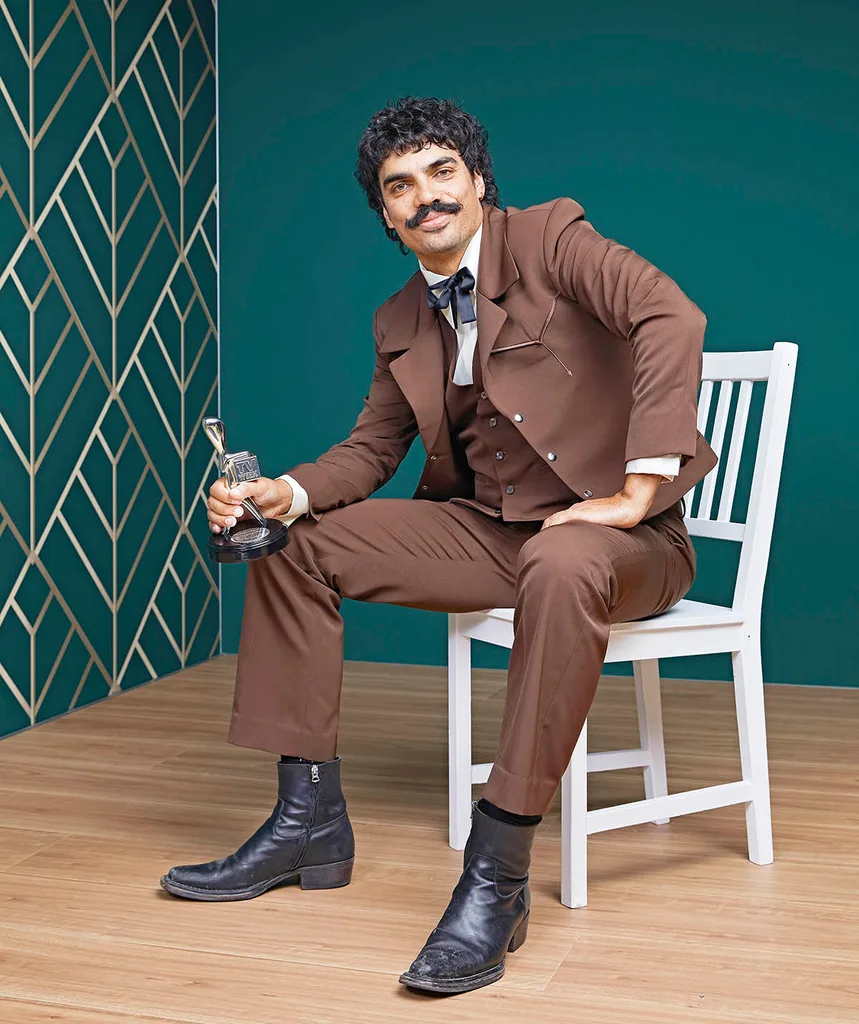 Tony Armstrong sits on a white chair wearing a brown suit and a smile as he clutches his Silver Logie, that he just won.