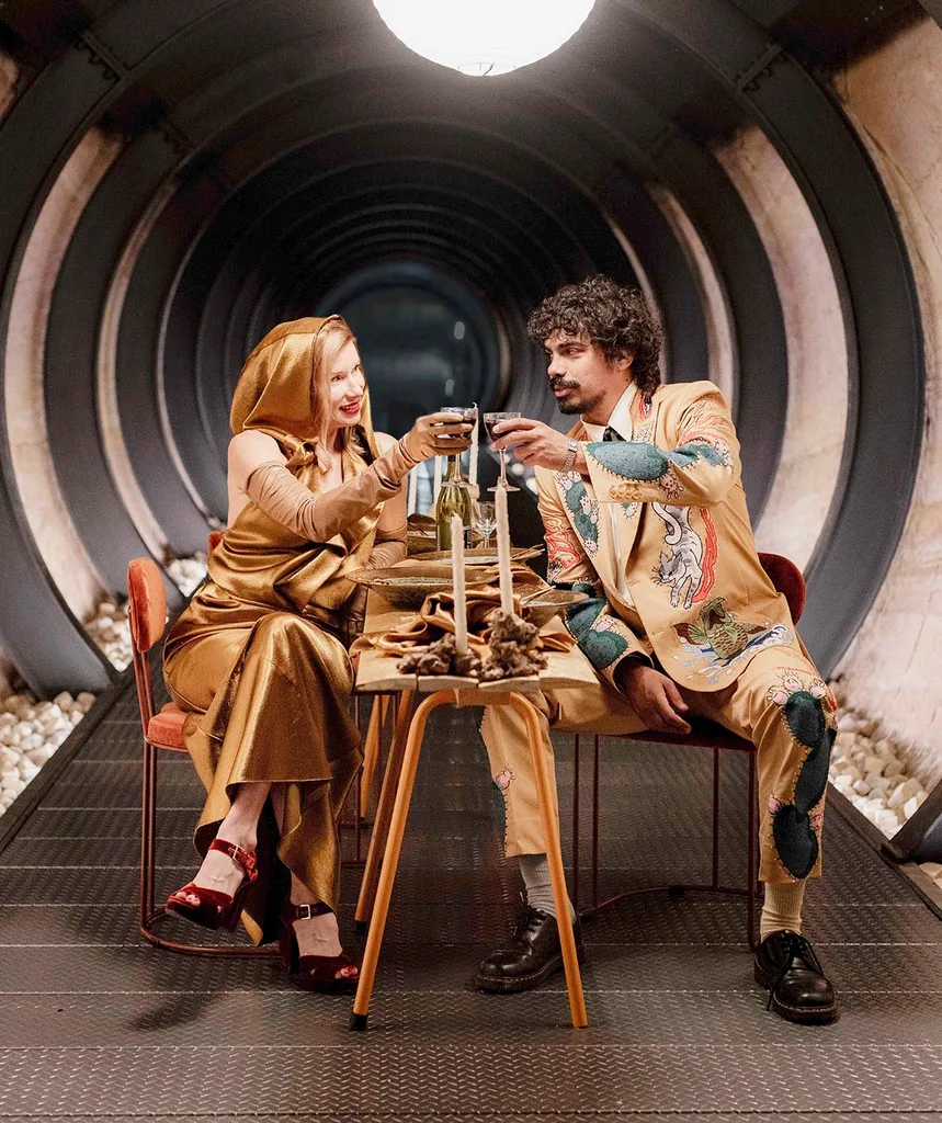 Kirsha and Tony cheers over a curious banquet while Tony wears a beige suit with animals printed on it she wears a orange silk dress