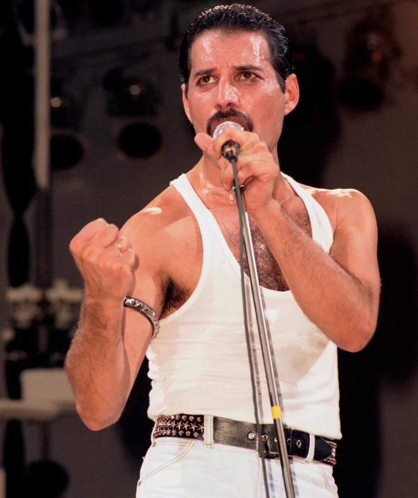 Freddie performs on stage in a white singlet and trousers
