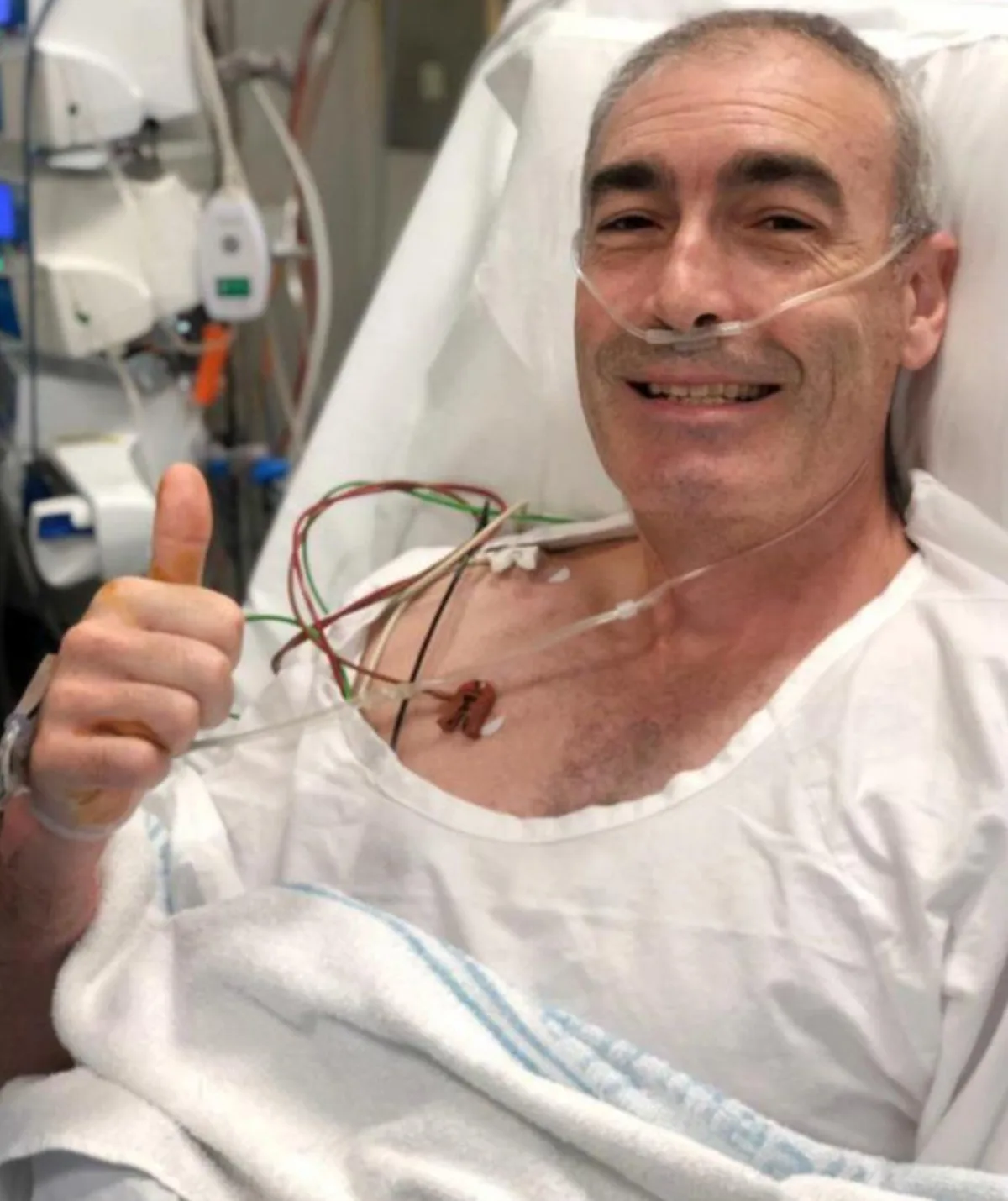 A man lies in a hospital bed giving the thumbs up sign.