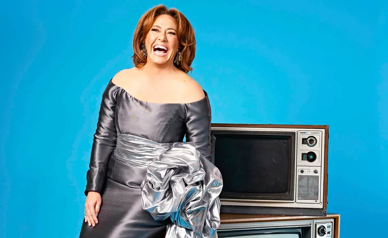 Lisa Millar, in a silver dress, poses next to a retro TV set, with a big smile on her face.