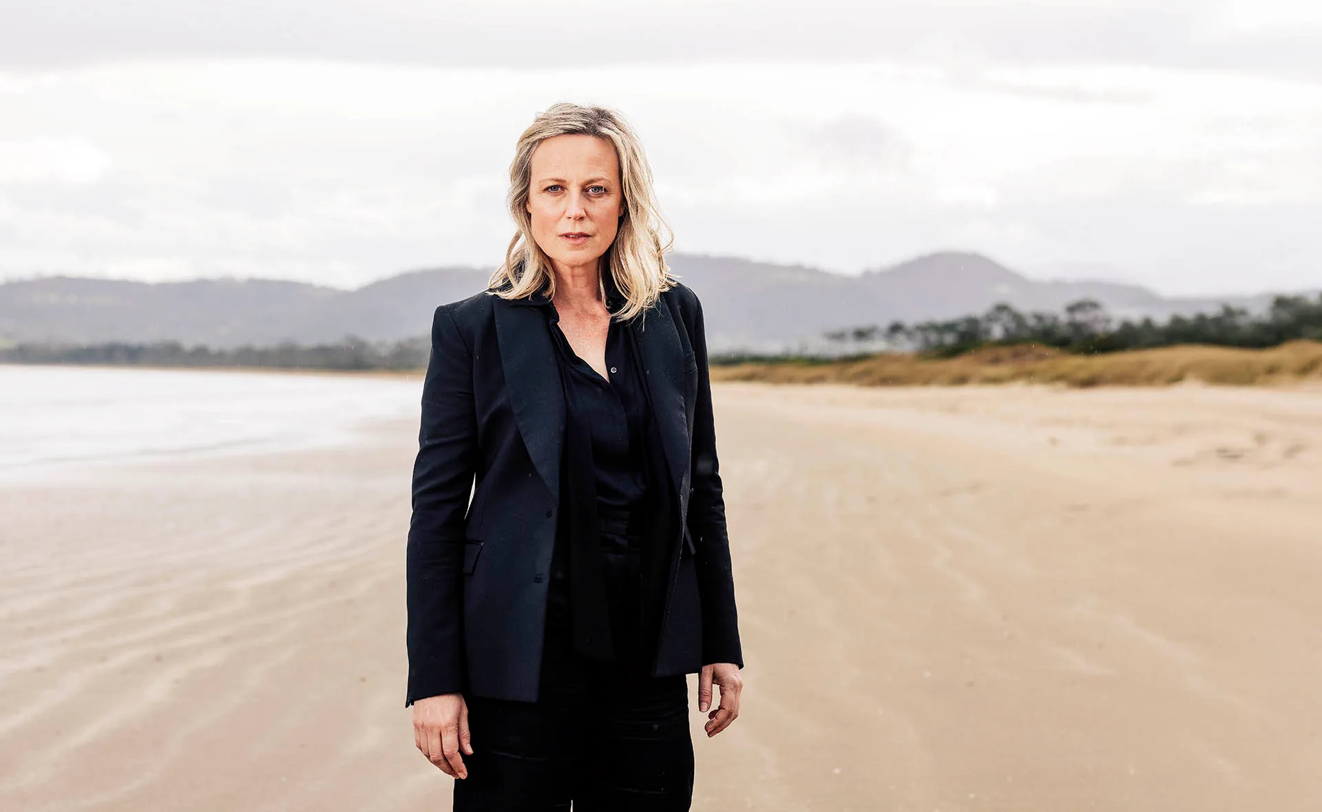 Marta Dusseldorp, in a black jacket, stands on a lonely beach