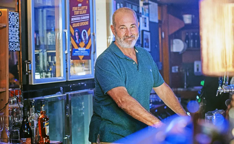 The Survivors. Martin Sacks as Julian in The Survivors. Pictured smiling behind a bar in a blue polo shirt.