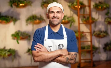 Theo is smiling with his arms crossed in a promo pic for MasterChef while wearing a blue polo shirt, white apron and white backwards cap