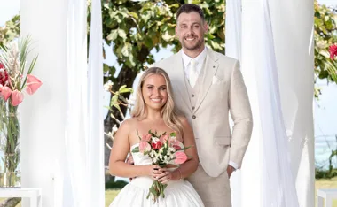 Amy left and Mike right pose in their wedding outfits, both smiling while Amy holds a bouquet of red, pink and white flowers. They stand surrounded by white drapes with a tree and ocean in the background.