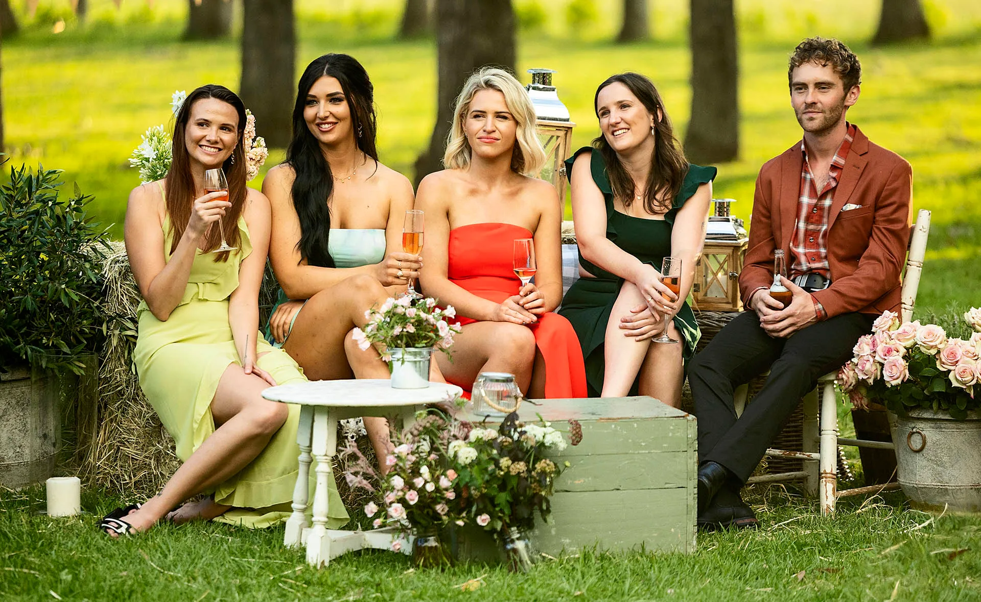 Farmer Jack (right) is pictured with four ladies of his farm all in beautiful gowns as they hold champagne glasses amongst a rustic setting of flowers, green grass, trees and little tables and chairs.