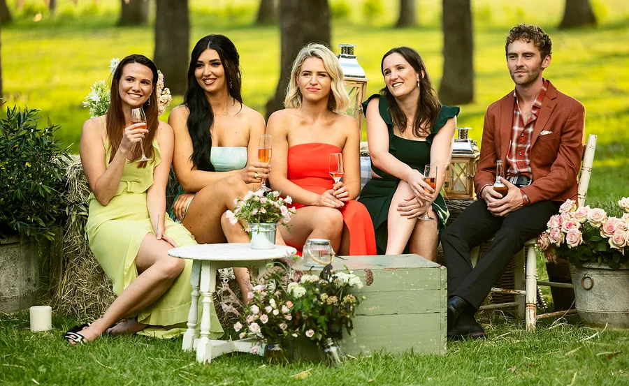 Farmer Jack (right) is pictured with four ladies of his farm all in beautiful gowns as they hold champagne glasses amongst a rustic setting of flowers, green grass, trees and little tables and chairs.