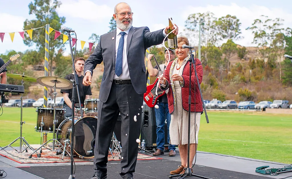 The Survivors. (L to R) Martin Sacks as Julian, Robyn Malcolm as Verity in The Survivors. Julian stands on stage in front of an older woman and a band while pouring champagne. The stage is set up on a green oval with a car park in the background.
