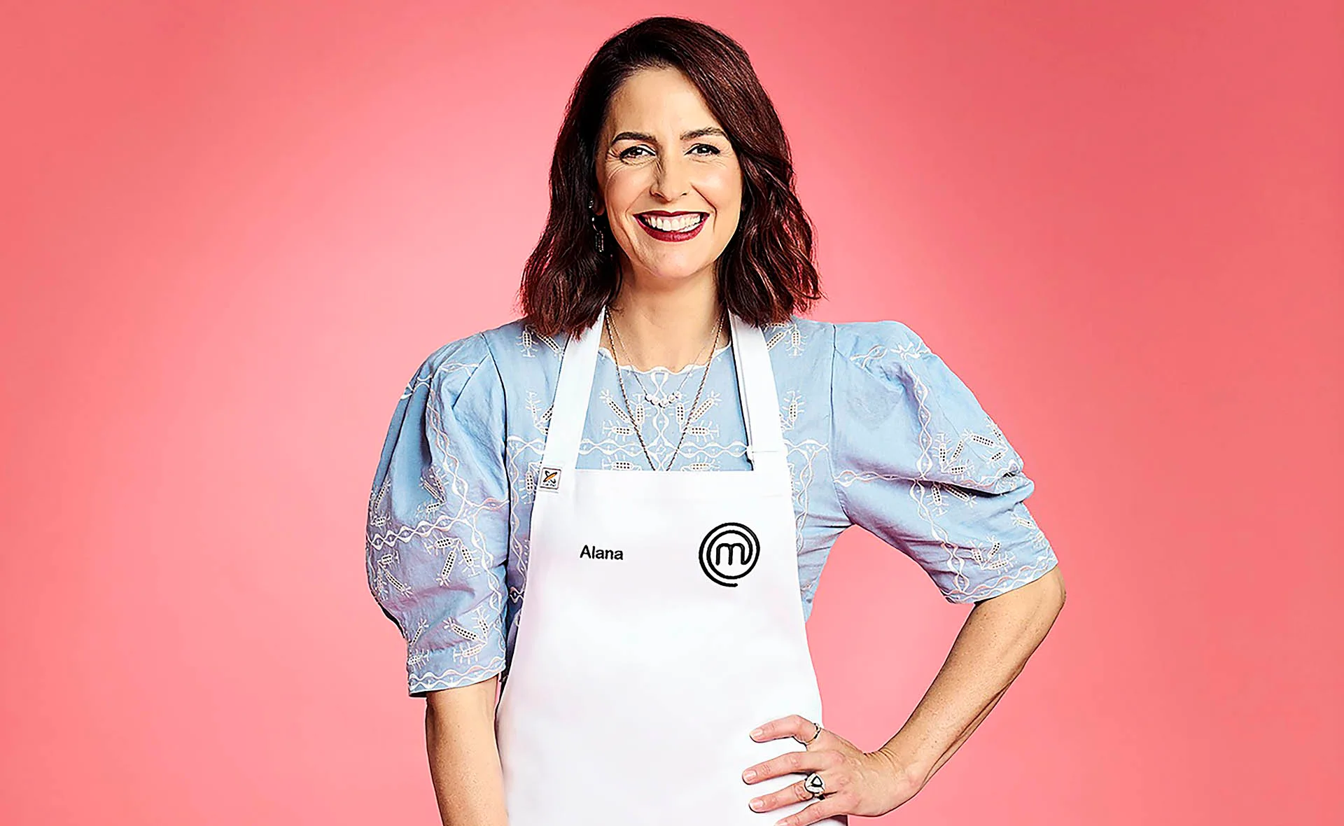 Alana Lowes smiling with her hand on her hip, wearing a blue top and white MasterChef apron. Pink background.
