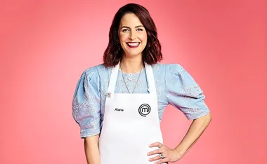 Alana Lowes smiling with her hand on her hip, wearing a blue top and white MasterChef apron. Pink background.