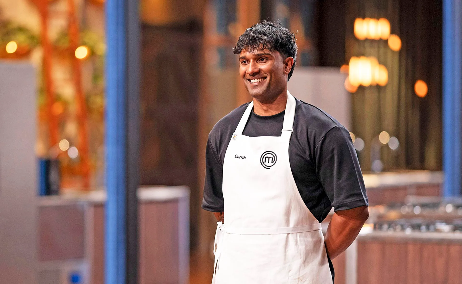 Darrsh in Back to Win kitchen smiling as he wears a black t-shirt and white MasterChef apron